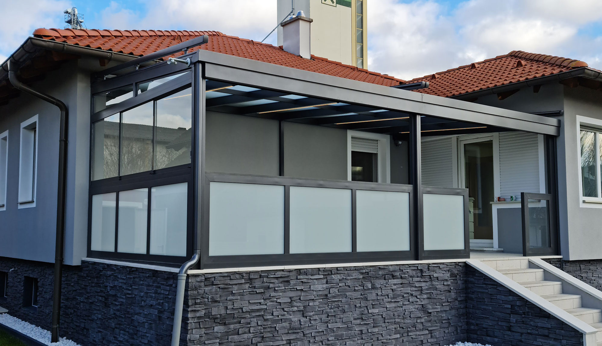 Terrassenüberdachung in anthrazit mit Glaseindeckung. Die Überdachung ist an eine Hausecke angebaut. Vorne ist eine halbhohe Absturzsicherung mit matten Glas, die linke Seite ist zusätzlich mit transparenten Glasschiebeelementen verbaut.