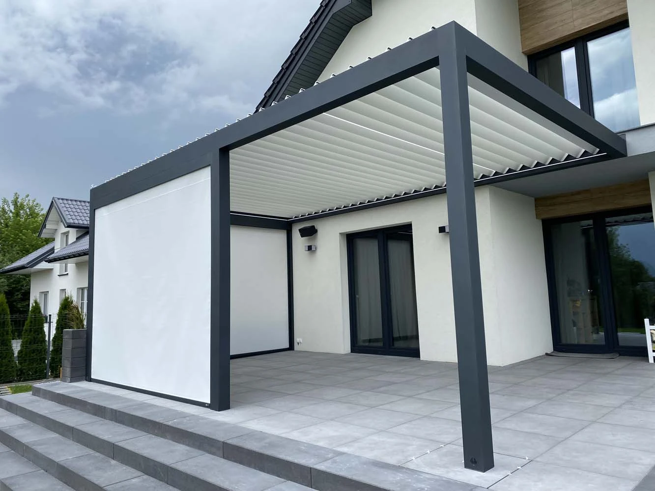 Auf dem Foto ist ein Lamellendach in anthrazit mit weißen Lamellen und weißen Senkrechtmarkisen zu sehen.